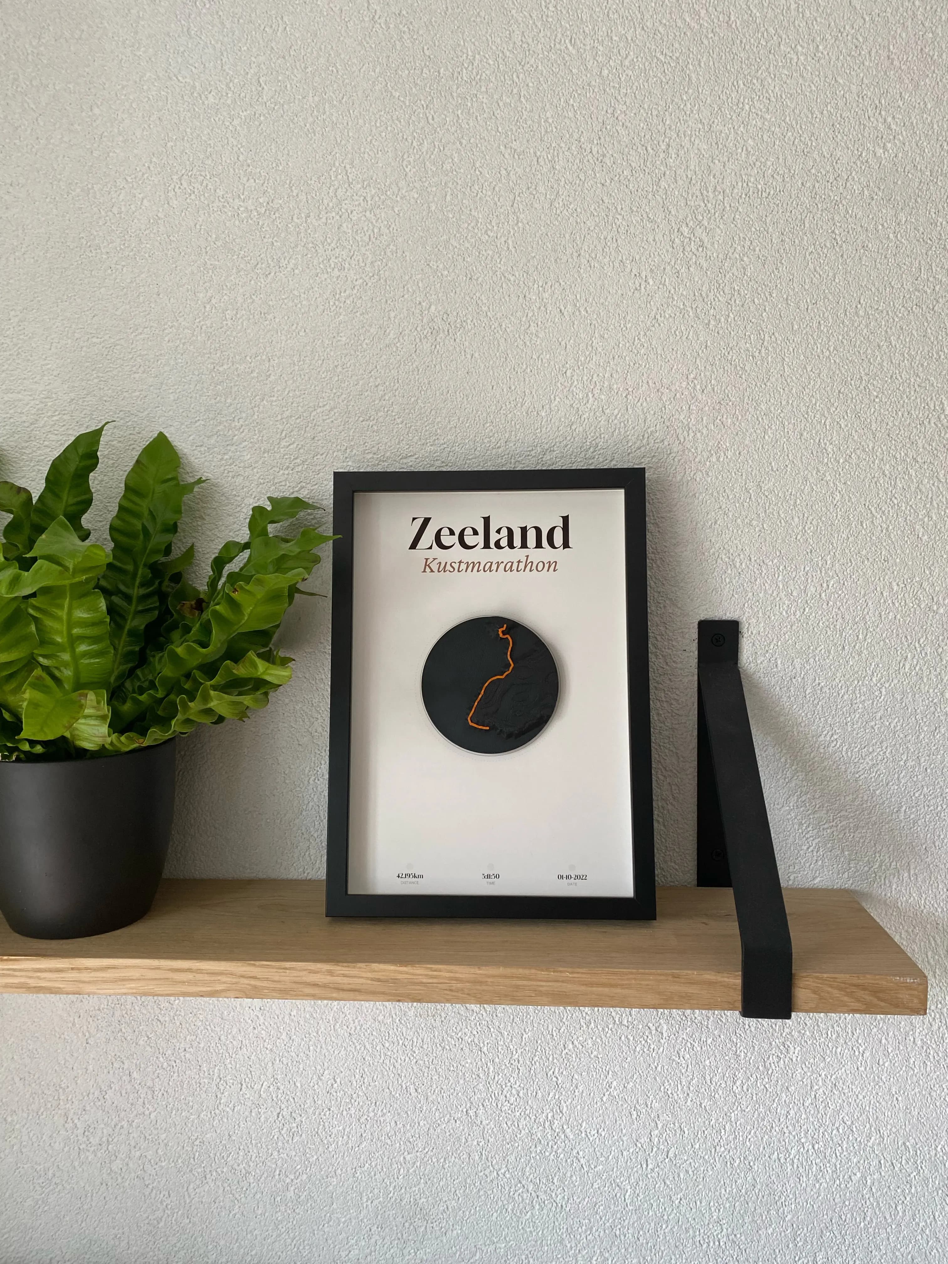 Ingelijste 3D-print van de Zeeland Kustmarathon op een houten plank naast een plant